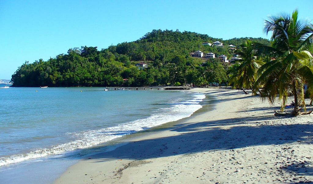 A serene beach with gentle waves lapping at the sandy shoreline, bordered by lush green hills and palm trees. A few buildings are nestled among the trees in the distance under a clear blue sky.