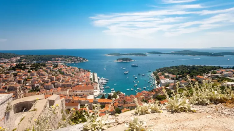 Aerial view of a coastal town with terracotta-roofed buildings and narrow streets leading to a picturesque harbor filled with boats. The turquoise sea is dotted with small green islands under a clear blue sky with wispy clouds.